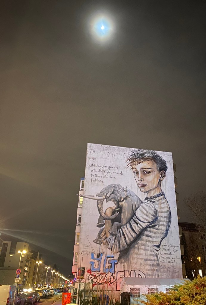 Massive street art on the gable of a large building. Youg boy holding an elephant. Text states “As long as you are standing, give a hand to those who have fallen.