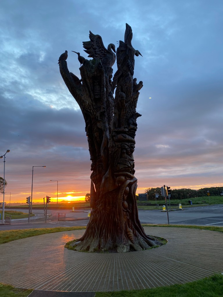 Dead tree with intricate wood carvings of Irish animals with the sun rising behind it over Howth