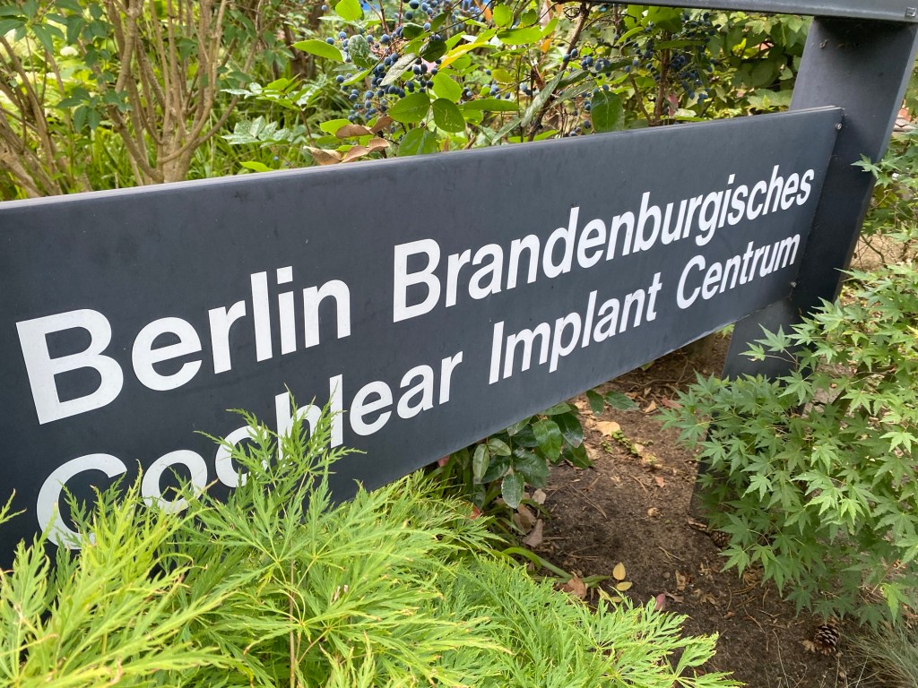 Sign for Berlin Cochlear Implant Centre surrounded by green plants. 