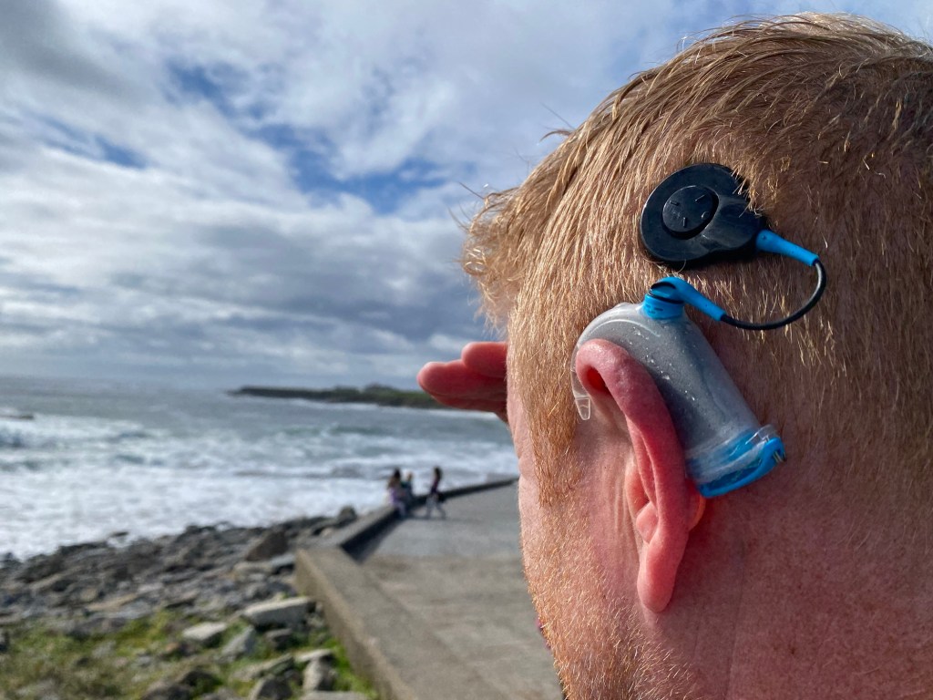 Man with short red hair wearing a Cochlear Aquakit looking out to sea after surfing. 