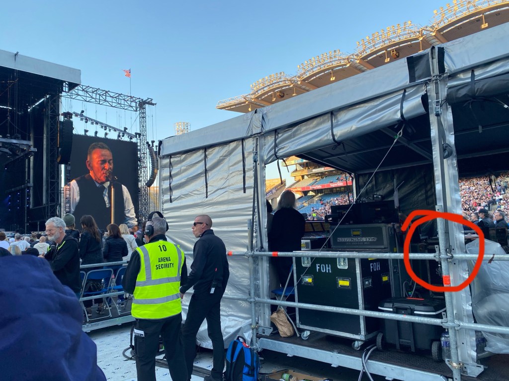 View of the side of the front of house unit at a Bruce Springsteen gig. A red circle highlights where a Minimic 2+ is hanging. Bruce Springsteen is on a large screen at the side of the stage.  