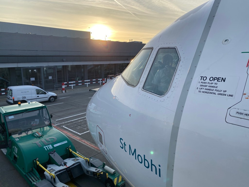 Sunrise over Dublin Airport. A green and white Aer Lingus plane is in the foreground. St. Mobhi is painted under the pilot’s windscreen. 