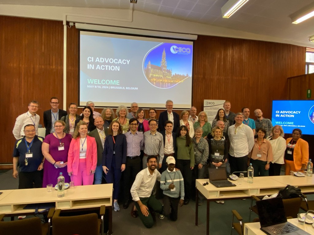 Group of nearly 40 Cochlear Implant recipients posing in front of a screen that reads “CI Advocacy in Action”