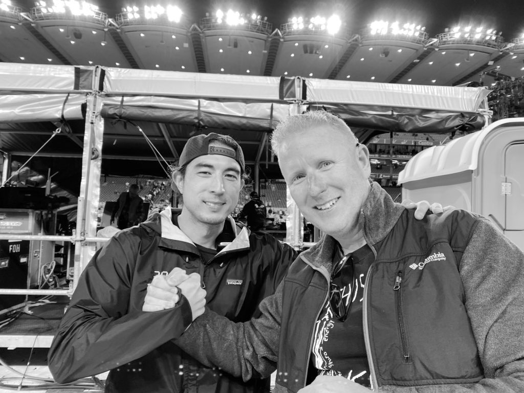 Two men shaking hands in front of a sound desk. Stadium lights glare behind them. 