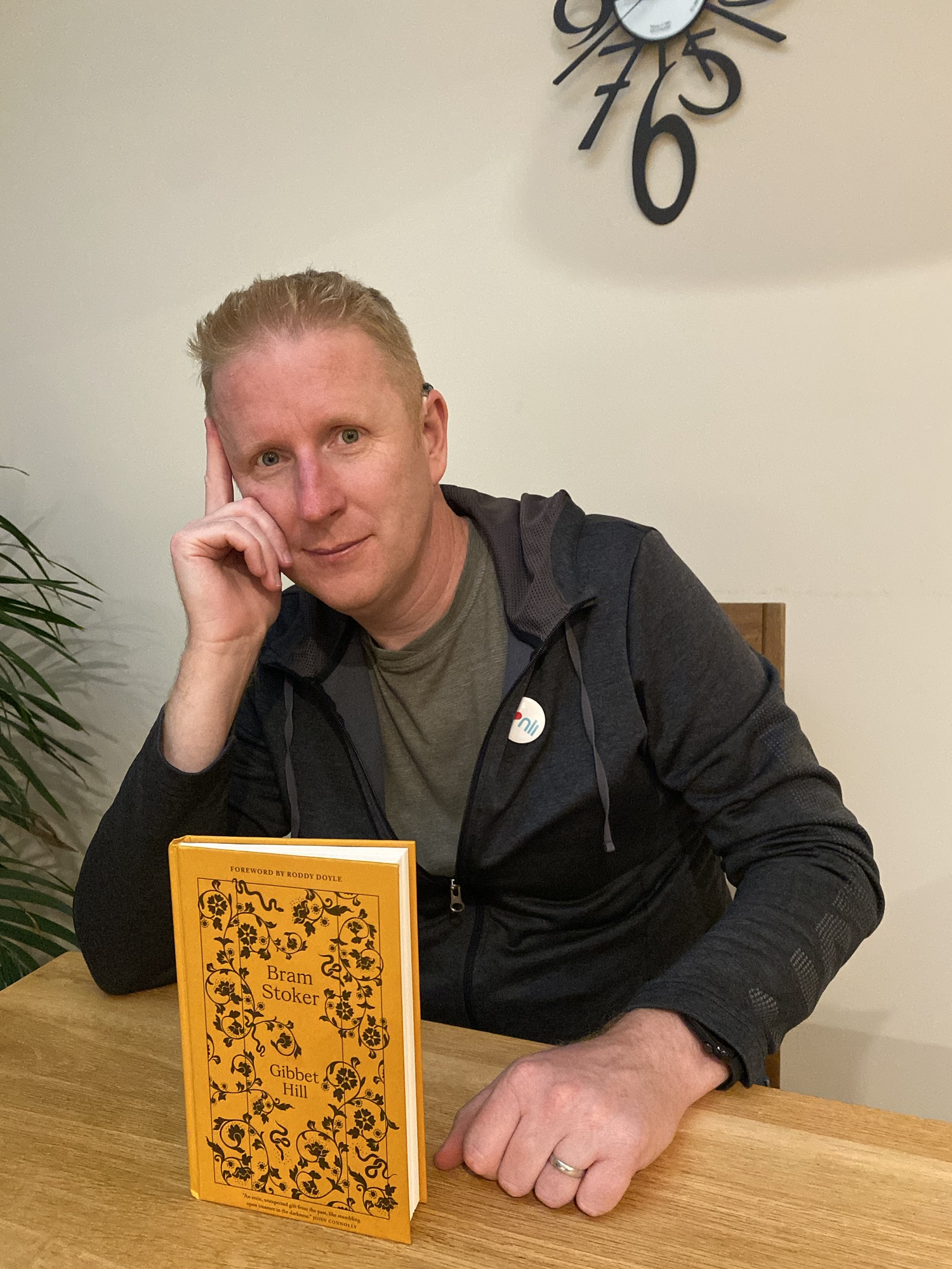 Man sitting at a table wearing a dark grey hoodie and green T-shirt. He wears a badge that says I❤️NLI (national Library of Ireland). On the table in front of him is a book by Bram Stoker called Gibbet Hill. Foreword by Roddy Doyle. "An eerie, unexpected gift from the past, like stumbling upon treasure in the darkness." JOHN CONNOLLY