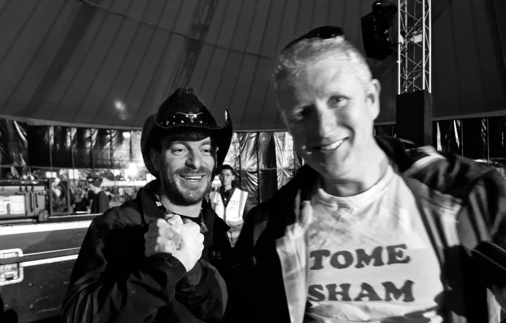 Two men smile and clasp hands in a concert tent with rigging and a sound desk behind them. They’re smiling. One wears a white t-Shirt with black lettering. “Tome Sham”